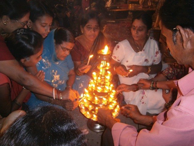 आरती में प्रयुक्त होने के लिए विशाल द्वीप प्रज्जवलित करने में सहायता करके पुण्य कमाते आम श्रद्धालु 